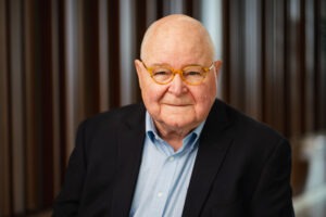 James Holbrook, a bald, older white man wearing glasses, a light-blue button-up shirt, and a navy blazer