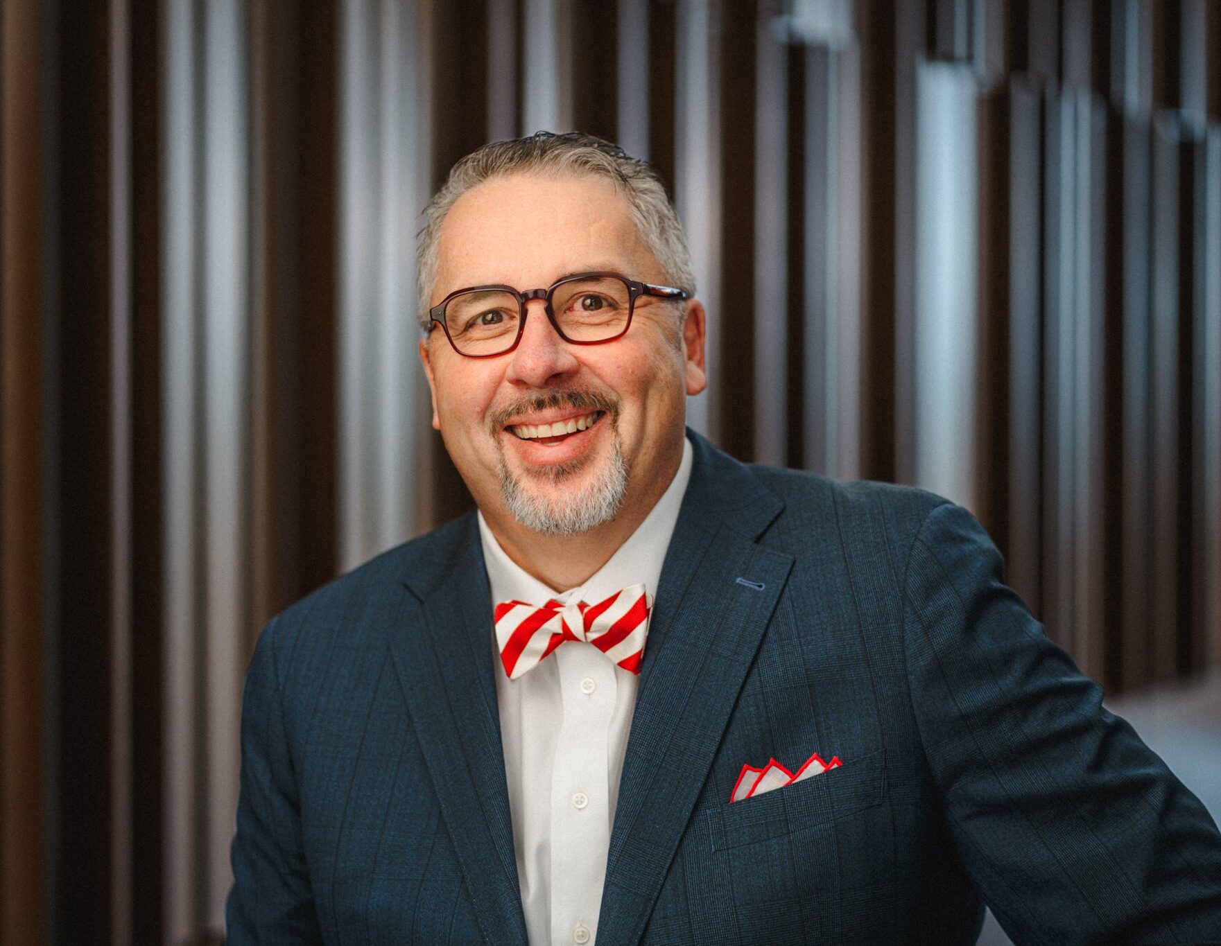 Arturo Thompson, a middle-aged man with greying hair wearing dark-rimmed glasses and a dark suit over a red and white striped bow tie.