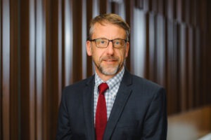 Professor John Ruple, a middle-aged white man with dark blonde hair, and short grey beard with dark spectacles