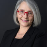 Professor Laura Kessler, a middle-aged woman wearing a dark blazer and round red rimmed glasses smiles for her picture