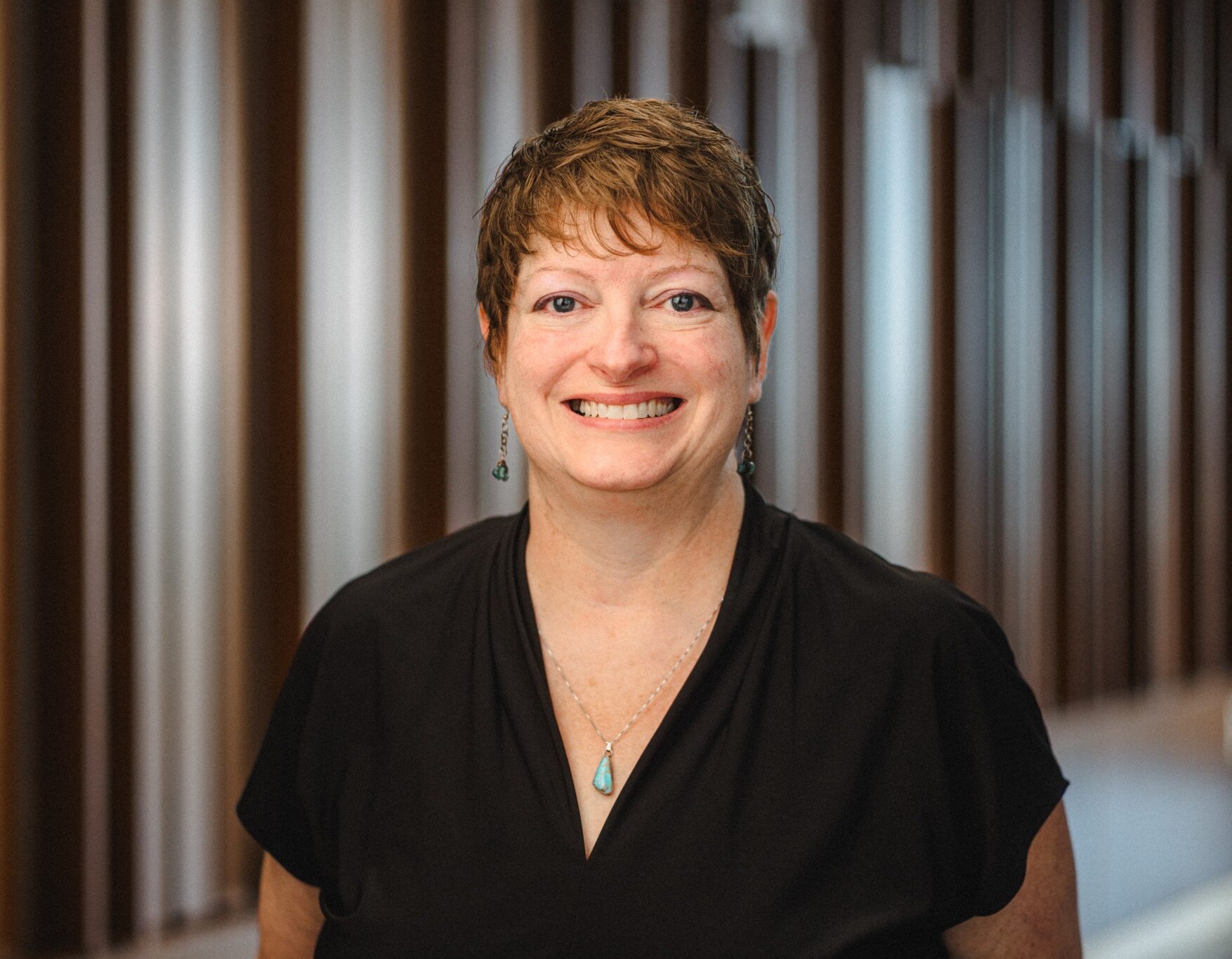 a middle-aged woman with short brown hair wearing a black blouse and a turquoise stone necklace.