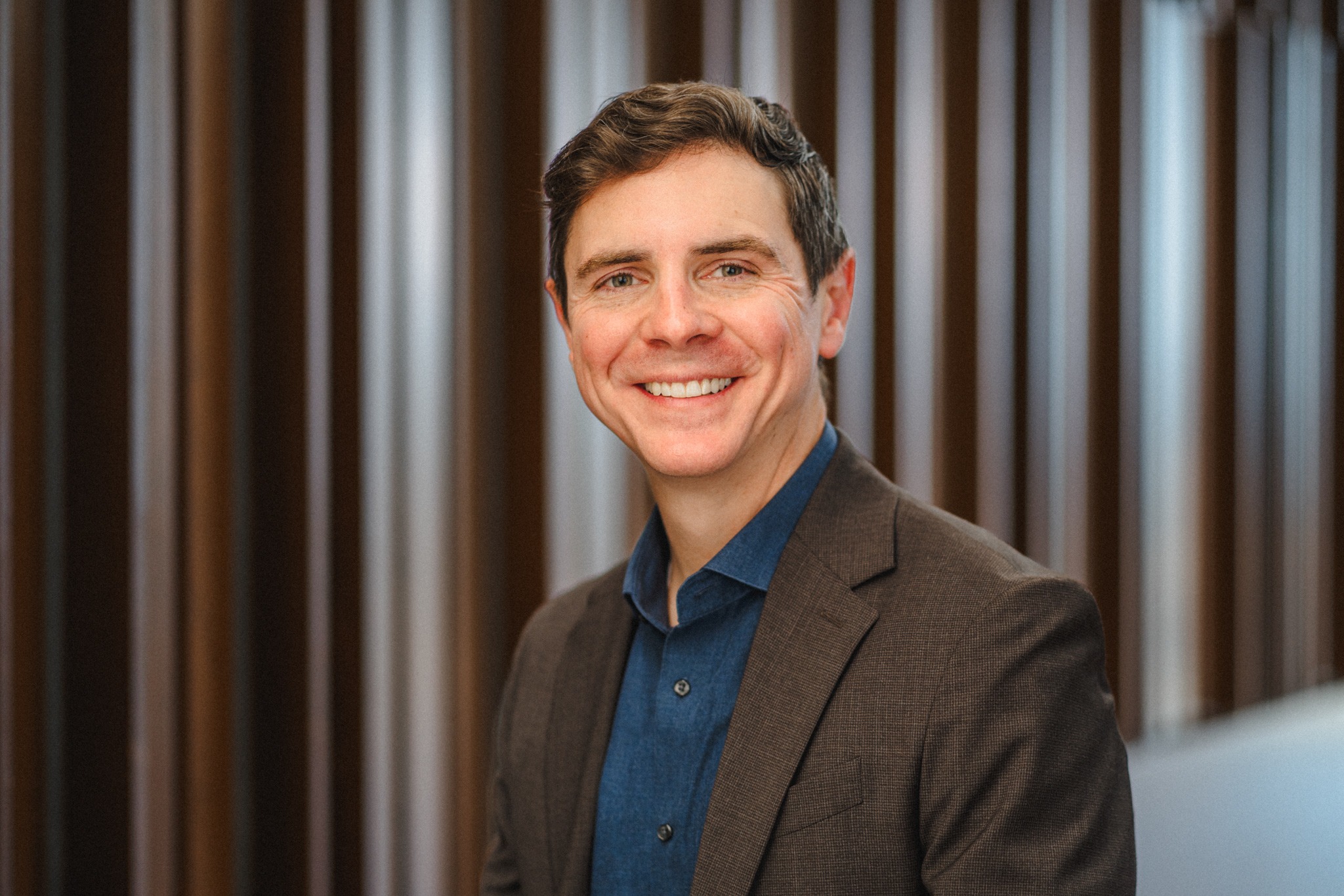 a middle-aged man with short brown hair wearing a brown blazer over a dark blue collared shirt