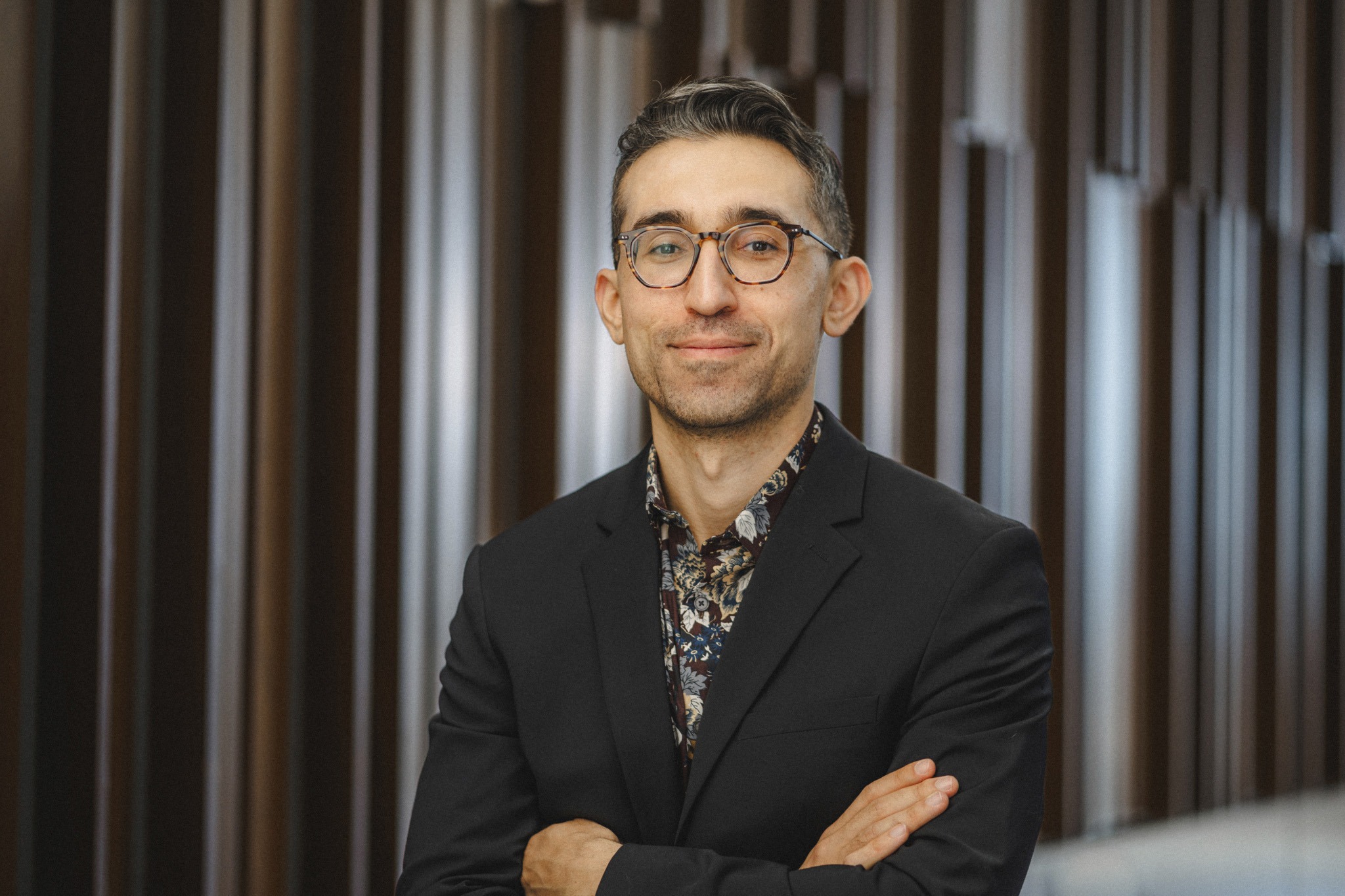 Associate Professor Daniel Aaron, a young man with olive skin and dark brown hair and a beard wearing a dark blazer and floral shirt