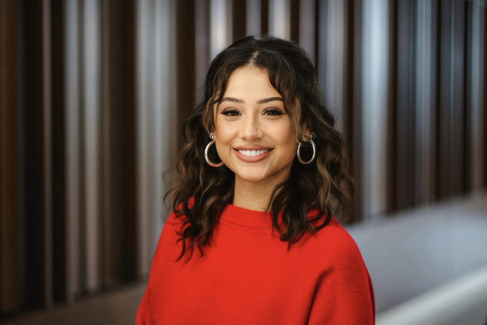 Amina Naveed, a young woman with dark hair and brown eyes wearing large hoop earrings and a red sweater