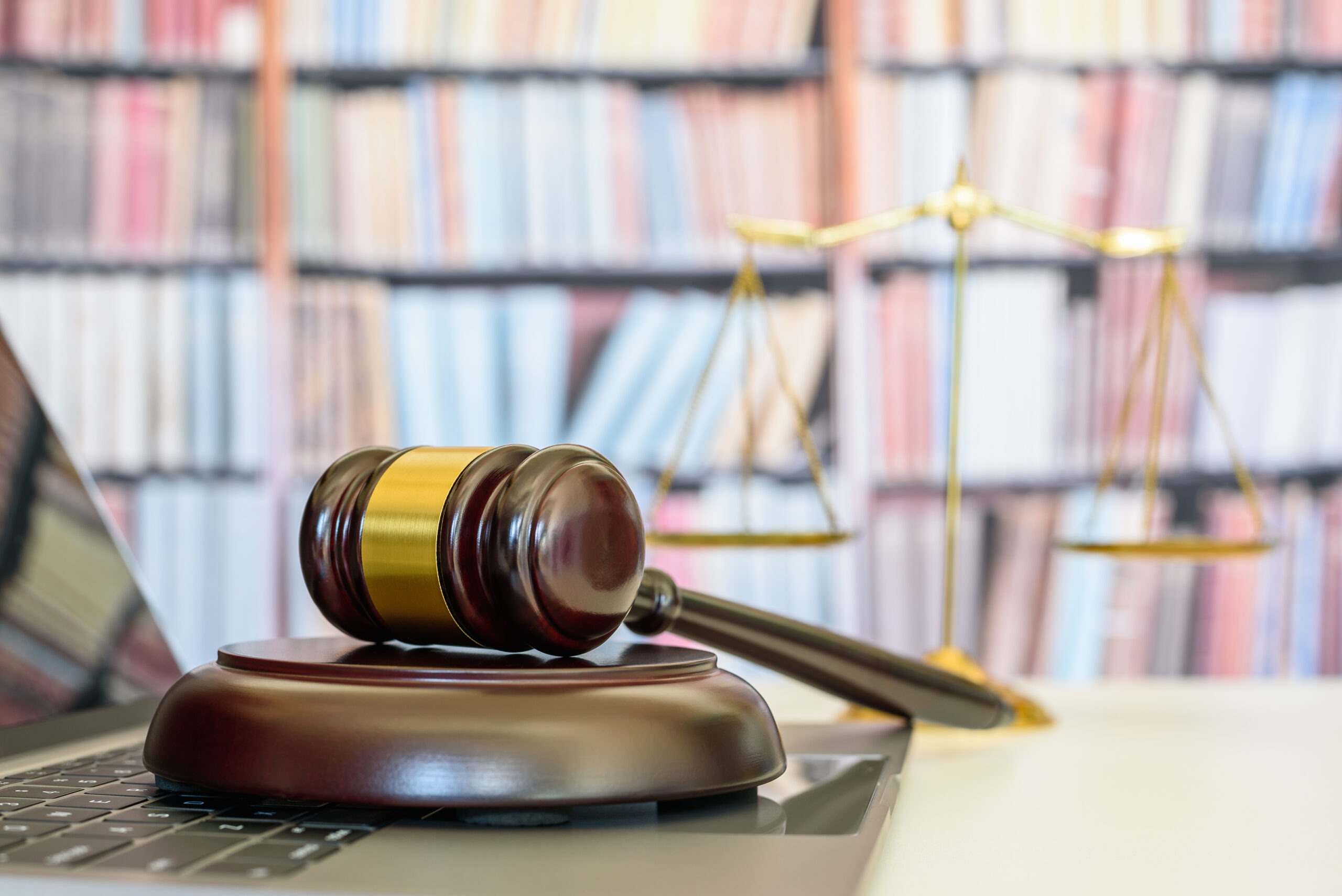 A laptop resting in front of a wall of books adjacent to a gold legal scale and brown wooden gavel with plate