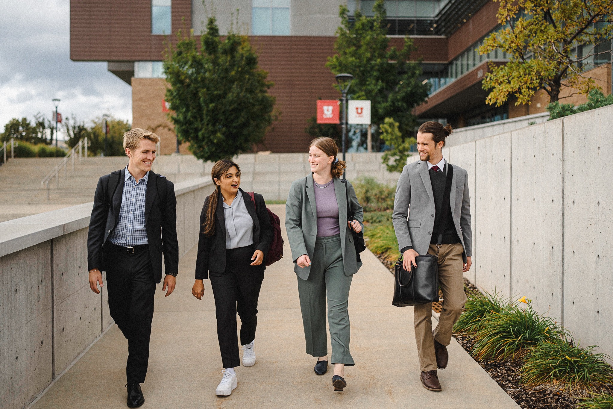 Four young law students (two men and two women) dressed in blazers and dress pants walk outside the College of Law beneath red and white flags that say "U Law" on them