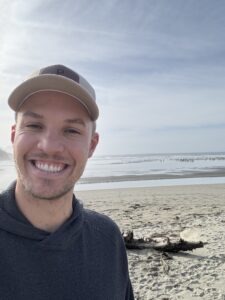 Mark Pittman, a young white man wearing a baseball cap and hoodie with a beach and the ocean in the background
