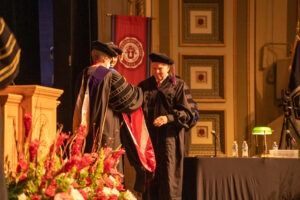 Dean Louisa Heiny hands F. Bryant McOmber, an older white man with short silver hair, a red sash during convocation in Kingsbury Hall