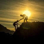 Two people helping each other up a mountain with a sunset behind them