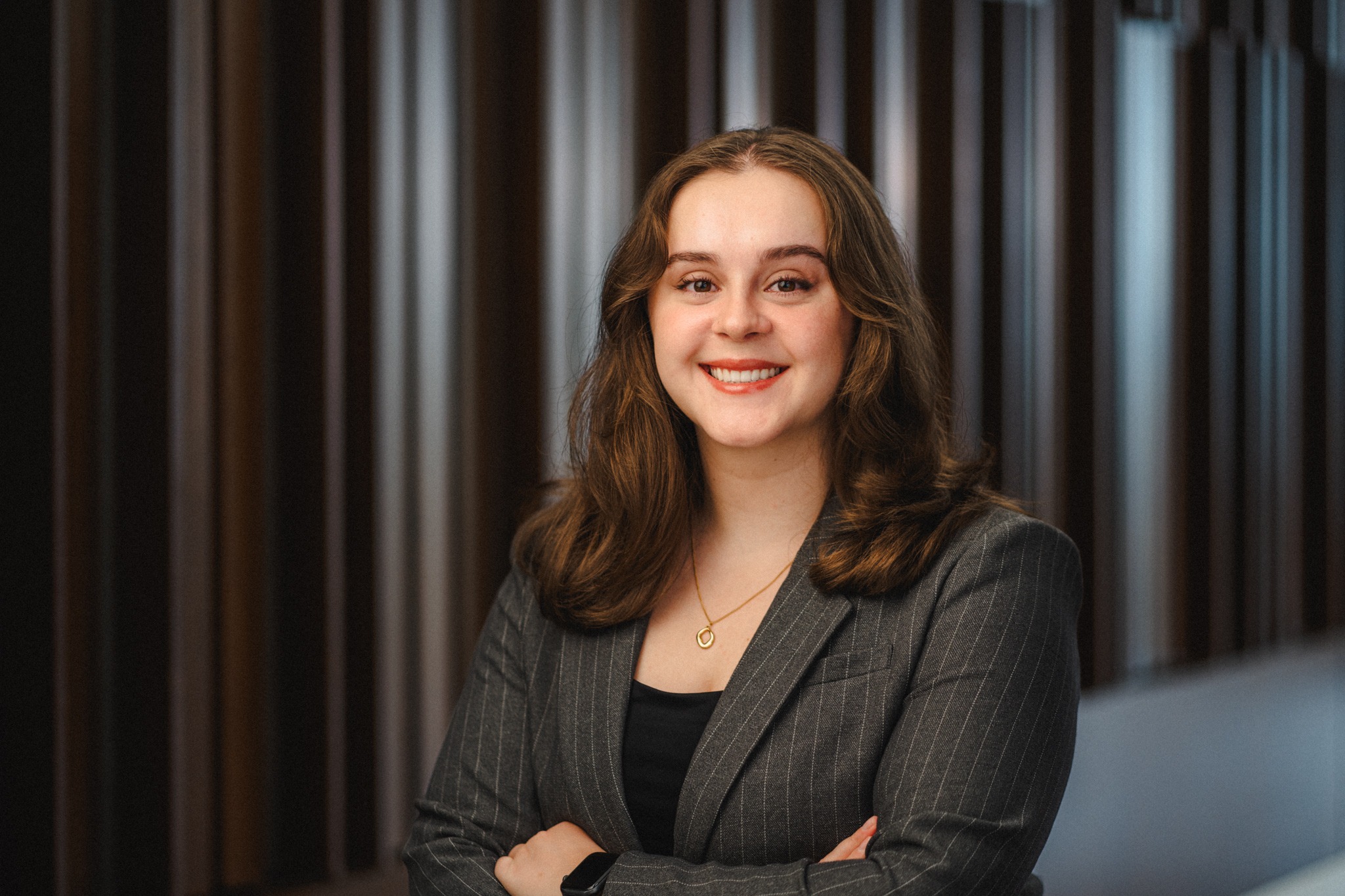 a young woman with long brown hair wearing a grey blazer over a dark shirt