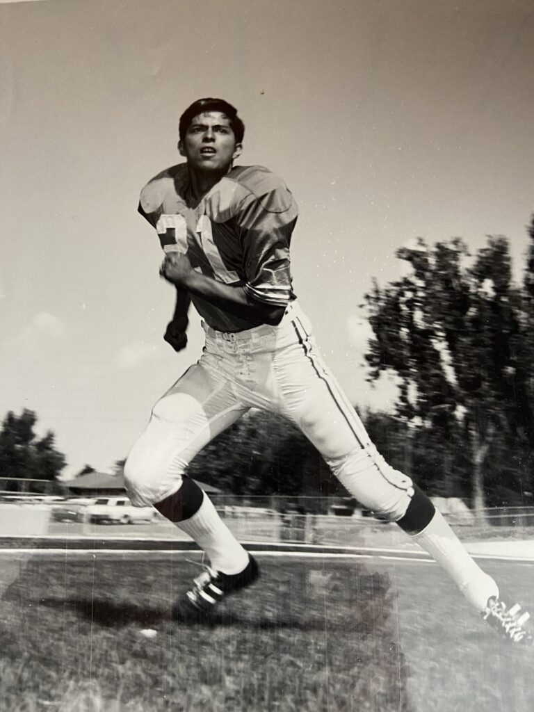 A black-and-white image of Larry EchoHawk, a Native American man with black hair wearing a football jersey and white athletic pants