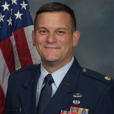 Jeff Baldridge, a young white man with short brown hair wearing a navy blue U.S. Airforce uniform and standing near a U.S. flag