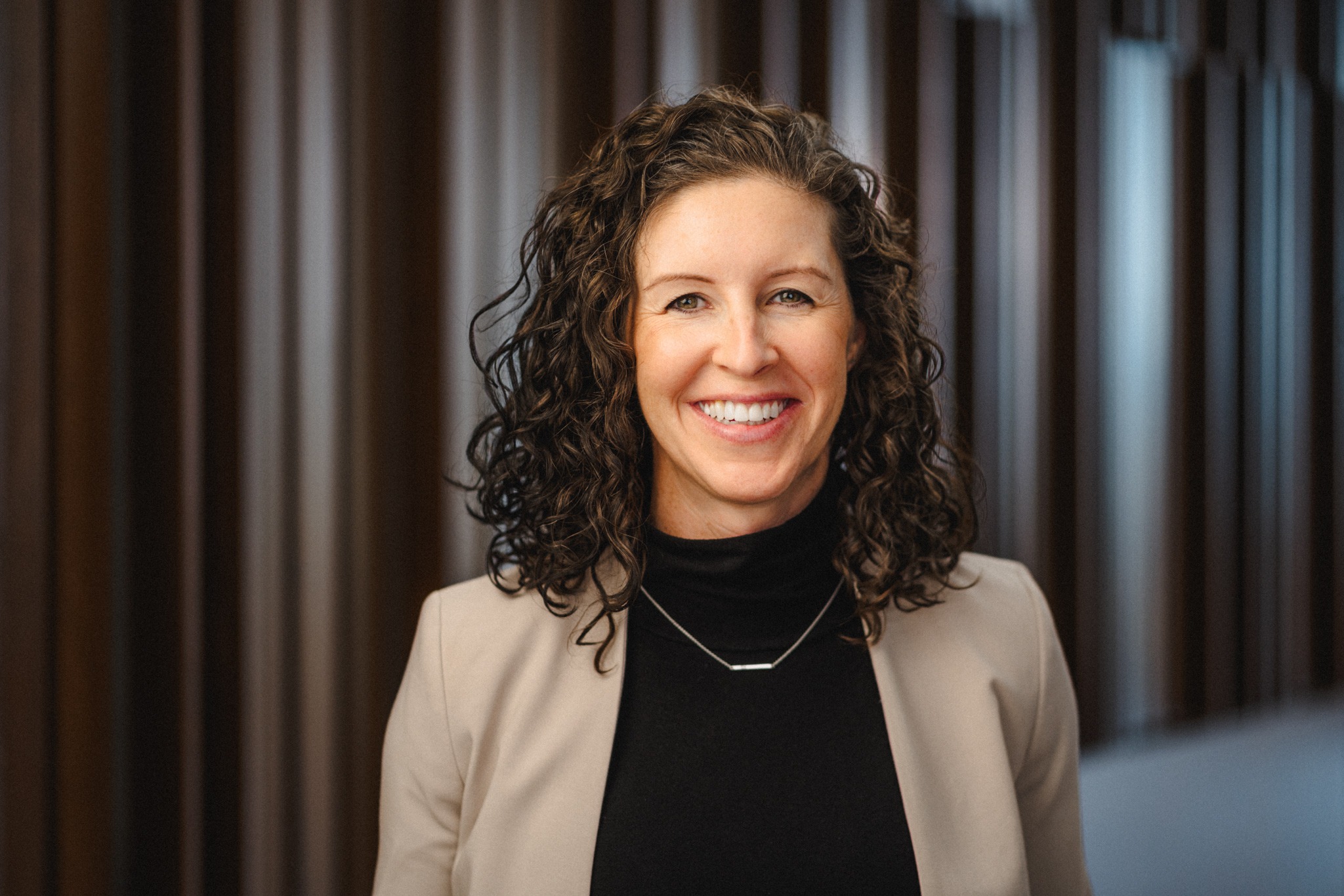 a middle-aged woman with long curly hair wearing a tan blazer over a black blouse