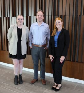 Lauren Harvey (left), a young woman with light blonde hair wearing a black dress and beige blazer; Landon Evans, a young white man with short brown hair wearing a light-blue collared shirt and dress pants, and Kate Menlove, a young white woman with long red hair wearing a black suit with a blue blouse