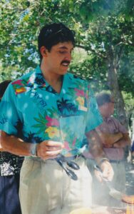 Dean Reyes Aguilar, a young Hispanic man wearing a teal shirt with Hawaiian flowers on it, grills burgers outside