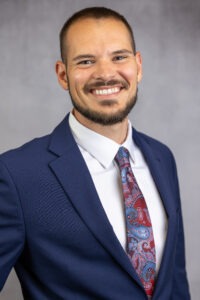 Brock Smith, a young Native man with short brown hair, brown eyes, and a beard wearing a navy blue suit