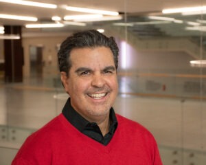 a middle-aged man with dark hair wearing a red v-neck sweater over a black collared shirt