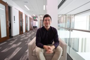 Nate Bramhall, a young half-Japanese man with short brown hair and brown eyes wearing a dark purple plaid button-up and khaki pants sitting opposite the law review offices in the College of Law building