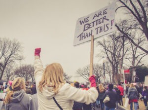 Crowd of people outside on a snowy day with a blonde-haired woman wearing a white coat and red gloves with her fist in the air and holding a sign that says "We are better than this!"