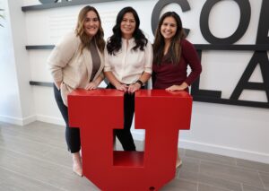 Three women (one with light-brown hair, one with black hair, and one with dark-brown hair) stand behind a red block letter "U" in the law building