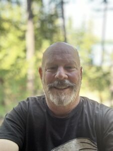 Mike Hanson, a middle-aged bald white man with a silver goatee sitting outside with trees in the background