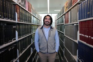 Austin Edens, a young man with olive skin, collar-length dark-brown hair and brown eyes wearing a blue button-up shirt, grey fleece vest and khaki pants, stands between two stacks of books in the law school library