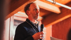 Steve Bloch, a middle-aged white man with short curly brown hair wearing a blue pullover sweater and holding a microphone