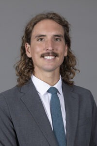 Matt Petersen, a young white man with shoulder-length curly brown hair, brown eyes and a brown mustache wearing a grey suit