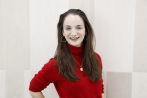 Alessandra Coote, a young white woman with long dark-brown hair and brown eyes wearing a red turtleneck sweater and a gold necklace