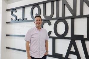 Parker Airmet, a young white man with short brown hair and brown eyes wearing a light pink button-up shirt and standing in front of a wall with black letters that say "S.J. Quinney College of Law"