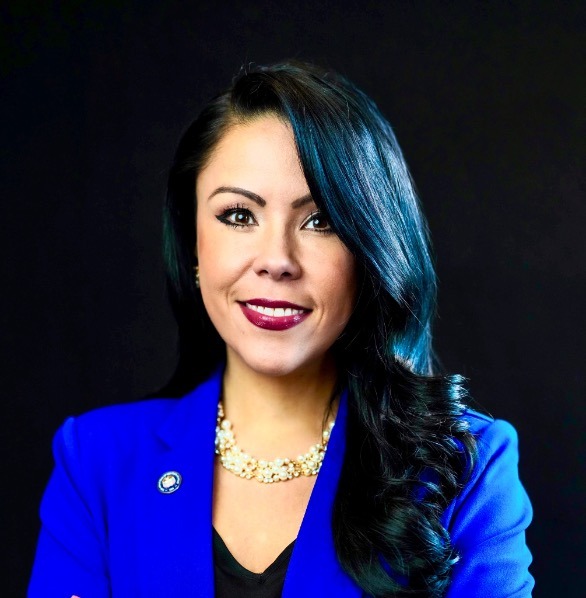 Nubia Peña, a young Latina woman with long black hair and brown eyes wearing red lipstick, a cobalt blue blazer, and a beaded pearl necklace