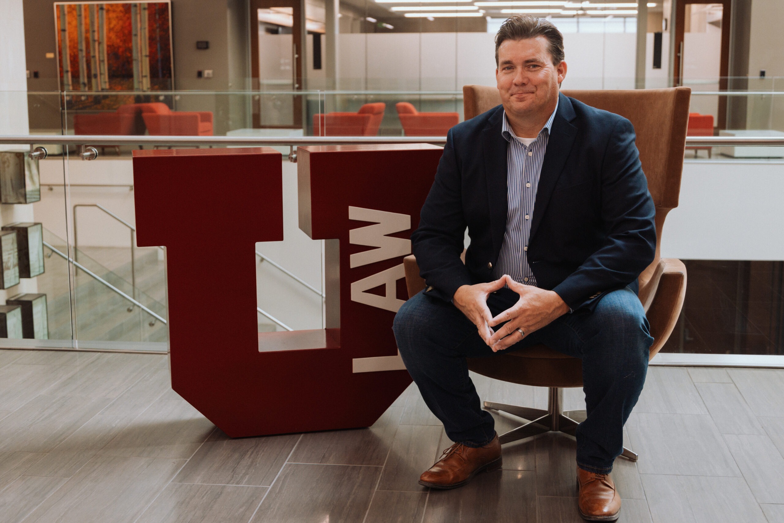 Travis Goff, a middle-aged white man with short dark-brown hair, sits next to the red block "U" in the College of Law wearing a light-blue button-up shirt, navy-blue blazer and jeans