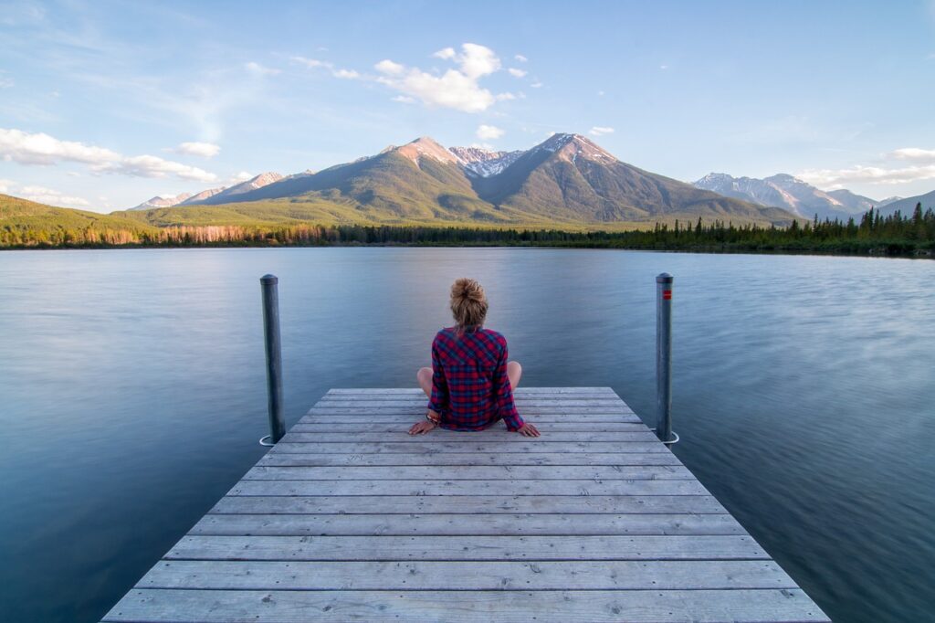 Woman sitting on a wooden dock jetting out into the middle of a clear blue mountain lake with a green mountain in the distance