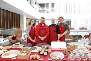 A group of young white people (two men and two women) wearing red Utah Law aprons stand behind a large table covered in baked goods during the Pro Bono Initiative bake sale