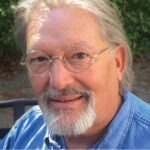 a senior man with white hair and goatee wearing spectacles and a denim collared shirt