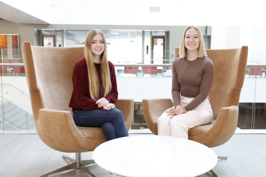 Two young white woman, both with blonde hair, sit in tan armchairs in the Utah Law building